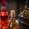 Three small bottles of liquor are arranged on a wooden surface. The bottle on the left is filled with red liquid and has a red cap. The bottle in the center is filled with amber liquid and has a gold cap. The bottle on the right is partially visible and al Produktbild-Vorschau