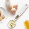 A digital spoon scale with a display showing 0.0, placed on a white surface with eggs, cookies, and pasta. Náhled obrázku produktu