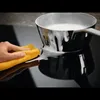 A hand is cleaning a stovetop with a yellow cloth, while a pot with foamy liquid is on the stovetop. Voir les images du produit