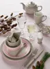 A table setting with a teapot, teacup, egg cup, cutlery, and decorative elements. Produktbild-Vorschau