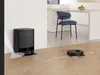 A black robot vacuum cleaner is shown cleaning a wooden floor in a modern kitchen setting, with a charging station and a chair visible. Produktbild-Vorschau