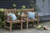 Two wooden garden benches are placed on a paved patio next to a dark wooden wall and some greenery. Cushions and a hat are placed on the benches. Produktbild-Vorschau