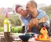 A couple embracing at a table set for a meal outdoors, with a vineyard in the background. Produktbild-Vorschau