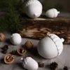 A collection of white ceramic acorn decorations and real acorns are arranged on a textured gray surface, with a piece of wood and pine needles in the background. Produktbild-Vorschau