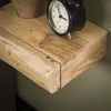 A close-up view of a wooden bedside table with a black alarm clock and a potted plant. Produktbild-Vorschau