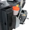 A close-up view of a part of a tire changing machine, showing a black storage bin, a white container with an orange lid, and a silver lever with a black handle. Produktbild-Vorschau