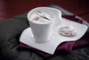 A white cup of coffee with foam and cinnamon, served with a cookie on a saucer, placed on a dark fabric surface. Produktbild-Vorschau