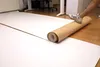 A person is unrolling a large roll of brown and white paper on a wooden floor in an office setting. Produktbild-Vorschau