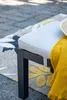 A close-up of an outdoor bench with a yellow blanket and a hat draped over it, placed on a patio with a patterned rug. Produktbild-Vorschau