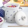 A white ceramic fermentation pot with blue floral accents, filled with shredded red cabbage and carrots, sits on a white surface next to its lid. A blue and white patterned cloth is visible to the left. Voir les images du produit