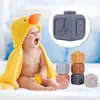 A happy baby wearing a yellow duck towel is sitting on a bed next to a stack of colorful rubber blocks. Voir les images du produit