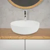 A white ceramic vessel sink is placed on a wooden countertop above white cabinets, with a concrete wall and a round mirror in the background. Náhled obrázku produktu