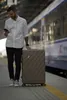 A man stands on a train platform next to a suitcase, looking at his phone. Produktbild-Vorschau