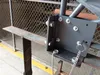 A close-up of a black metal bracket with bolts and chains attached, mounted on a metal railing. A wire mesh fence is visible in the background. Produktbild-Vorschau
