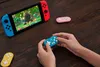 A Nintendo Switch console is on a wooden table, with a person holding a small turquoise gamepad. Other small gamepads are also visible. Náhľad obrázku produktu