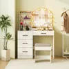 A white vanity table with a lighted mirror, drawers, and a stool, placed in a room with a plant and a clothing rack. Voir les images du produit