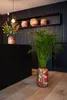 A decorative plant pot with a floral pattern sits on a wooden floor in front of a black counter. A bouquet of flowers is in a smaller pot on the counter. Produktbild-Vorschau