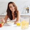 A woman is having breakfast in a kitchen, with a bowl of cereal, a glass of orange juice, and apples on the table. A cereal dispenser is also visible. Produktbild-Vorschau