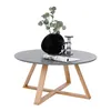A round coffee table with a gray top and wooden legs, decorated with a black vase containing greenery and two glass candle holders, and a rolled-up magazine. Náhled obrázku produktu