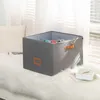 A gray fabric storage bin with brown handles and a label holder sits on a white fluffy rug next to a window with blinds. Náhled obrázku produktu