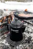 A black coffee pot sits on a grate over a campfire, with a cast iron pan cooking food in the background. Produktbild-Vorschau
