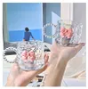 Two clear glass mugs with pink bows are held by hands in front of a painting of a person looking out at the sea. Produktbild-Vorschau