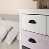 A close-up view of a white dresser with black handles, showing two drawers. The top drawer is white, and the drawer below it has a distressed pink wood grain texture. In the background, a blurred bed with white pillows is visible. Náhľad obrázku produktu
