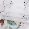 A close-up view of a white sheer curtain with black embroidered swirls and dots, layered over a white knitted fabric. A light blue teacup with a frothy beverage sits on a saucer, partially visible beneath the curtain. Produktbild-Vorschau
