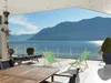 A patio overlooking a lake and mountains, with two green deck chairs and a wooden table. Produktbild-Vorschau