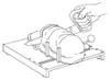 A hand is shown operating a device that appears to be slicing a large food item, possibly a ham or a large loaf of bread, on a cutting board with guides and a handle. Produktbild-Vorschau