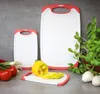 Three cutting boards of different sizes are arranged on a gray countertop, with some vegetables and herbs in the foreground and background. Anteprima dell’immagine prodotto