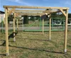 A wooden carport structure is set up outdoors on a grassy area, with a green fence and cars visible in the background. Produktbild-Vorschau