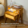 A wooden nightstand with two layers, featuring a woven cane panel on the lower drawer, placed next to a bed. Náhled obrázku produktu