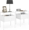 Two white bedside tables are shown against a white background. One table has a lamp, a framed picture, and a wallet on it. The other table has a digital clock, a book, and a cup on it. Náhled obrázku produktu