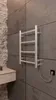 A silver electric towel warmer is mounted on a gray tiled wall in a bathroom. A small wooden shelf with a bottle is to the left. A coiled cord connects the warmer to a black electrical outlet. Produktbild-Vorschau