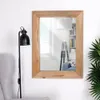 A mirror with a wooden frame is hanging on a white wall, next to a floor lamp and a chair. A plant is visible on the right side. Produktbild-Vorschau
