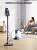 A woman and child are sitting on a couch in a living room, while a cordless vacuum cleaner stands upright in the foreground. Produktbild-Vorschau