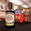 A bottle of gin is placed on a dark surface with a blurred background of a room and some decorative elements. Voir les images du produit