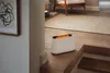 A white humidifier with a flame effect sits on a wooden floor next to stairs, in a home setting with a rug and chair in the background. Produktbild-Vorschau