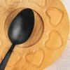 A close-up view of a black spoon resting on a wooden tray with heart-shaped indentations. Náhled obrázku produktu