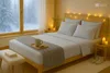 A cozy bedroom scene with a bed made up with a silver-gray fitted sheet, a knitted throw, and a mug of coffee on a tray. The room is lit by warm fairy lights and candles, with a snowy forest visible through the window. Produktbild-Vorschau