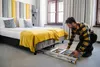 A man is kneeling on the floor next to a bed, pulling a folded aluminum ladder out from underneath it. The room appears to be a hotel room with two beds, a headboard, curtains, and a window. Anteprima dell’immagine prodotto