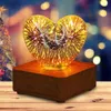 A heart-shaped glass lamp with a 3D firework effect sits on a wooden base, with a blurred Christmas tree and bokeh lights in the background. Produktbild-Vorschau