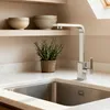 A modern kitchen sink area with a stainless steel faucet, a potted rosemary plant, and shelves with bowls in the background. Produktbild-Vorschau