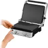 A close-up shot of a silver and black electric grill with two hands interacting with it. Podgląd zdjęcia produktu