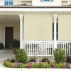 A beige outdoor roller blind is installed on a porch, providing shade and privacy. The blind is partially rolled down, covering the railing and a window. In front of the porch, there is a colorful flower bed. Voir les images du produit