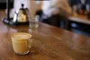A cup of coffee with latte art sits on a wooden table in a cafe, with a barista in the background. Náhľad obrázku produktu