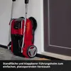 A red and black battery-powered sweeper is leaning against a wall next to a dark door. Produktbild-Vorschau