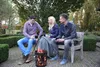 Three people sitting around a fire pit in a garden, holding mugs. Produktbild-Vorschau