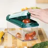 A hand is using a vegetable slicer to cut tomatoes over a clear container on a wooden cutting board. Voir les images du produit
